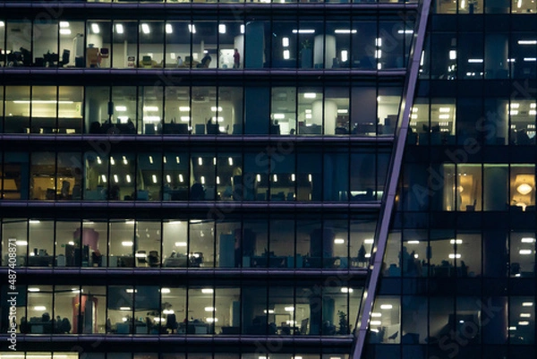 Fototapeta Workers in a box. Office building light at night. A skyscraper made of glass and metal lighr up in the evening. Windows of Skyscraper Business Office.