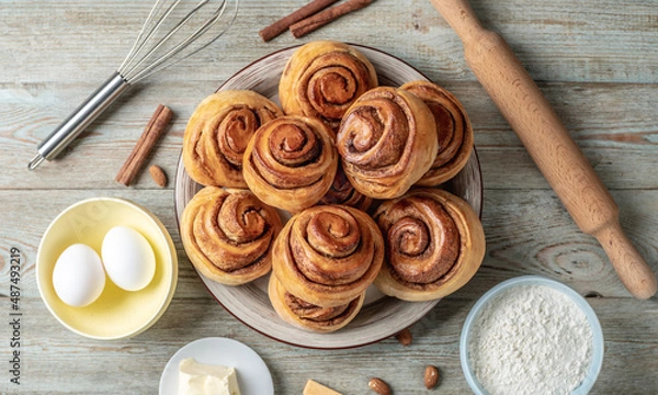 Obraz There is a plate with homemade fresh delicious cinnamon rolls, ingredients for their preparation and kitchen tools on the wooden table . Top view