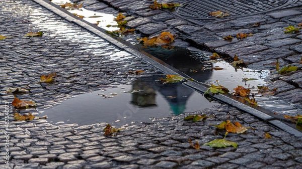 Obraz Puddle of water with a wet cobblestone floor, with the reflection of a building and full of autumn leaves.
