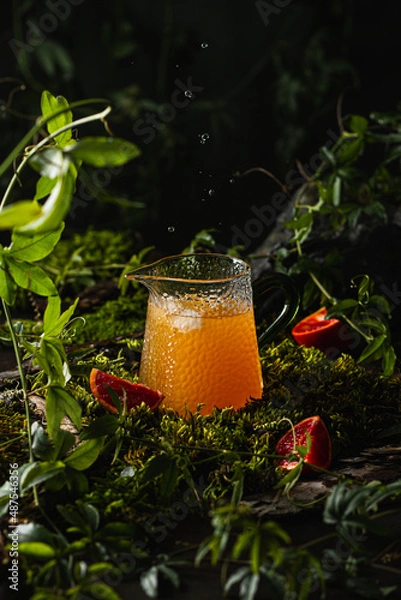 Fototapeta Fresh orange juice in a glass decanter and red orange fruit slices on green moss. Orange juice splash