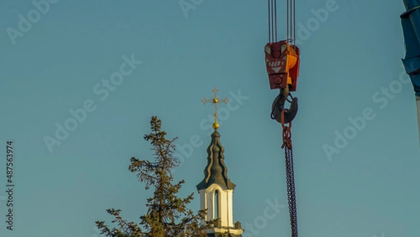 Fototapeta Red hook of a crane on the background of the tower church with orthodox cross. Construction site.