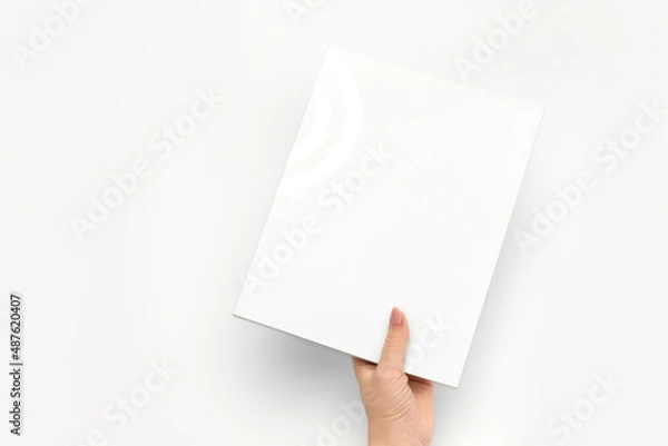 Fototapeta A woman's hand holds a closed book with a blank white cover on a white background.