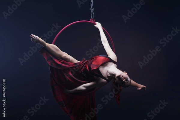 Obraz Circus performer woman in red dress doing tricks on red Lyra isolated on black background.
