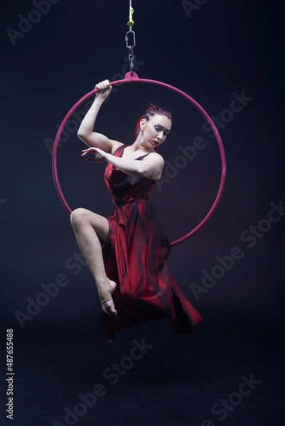 Obraz Circus performer woman in red dress doing tricks on red Lyra isolated on black background.