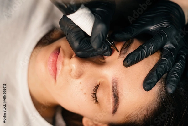 Obraz Beauty masters hands do permanent eyebrow makeup. Minimal trauma to skin. Eyebrow microblading is performed using manipulator handle and special nozzle with needles. Cosmetologist skill level