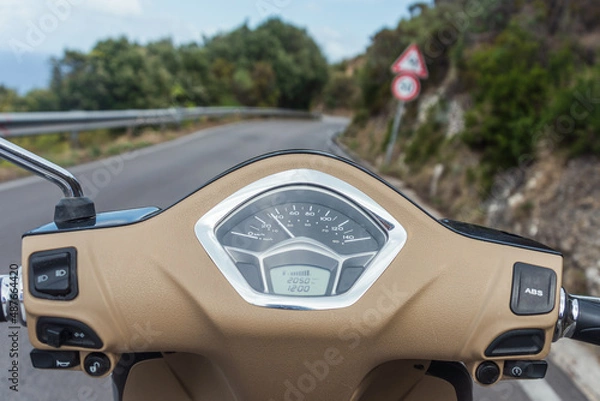 Obraz View of the winding road from behind the steering wheel of a scooter. Biker driving his motorcycle on country road in a village. Point of view with focus is on the handlebar
