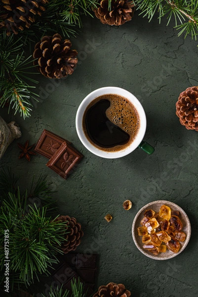 Fototapeta Black coffee in a green cup, chocolate and caramel sugar on a green background with fir branches and cones. Top view