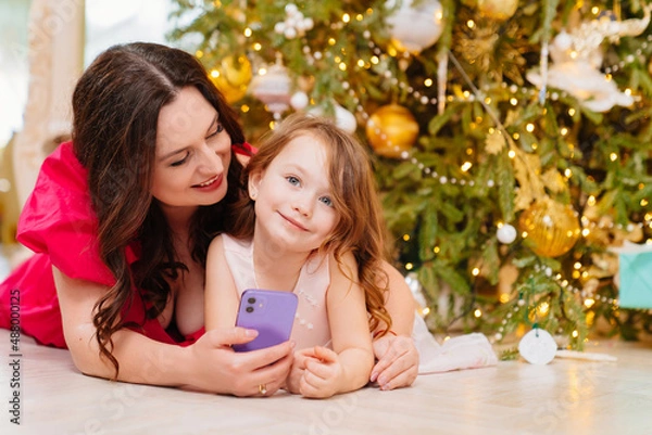Fototapeta mother and daughter under the Christmas tree with a smartphone.