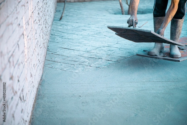 Obraz Worker stamping the drawing on the concrete around the pool, with tool in hand and wellies, preparing the garden