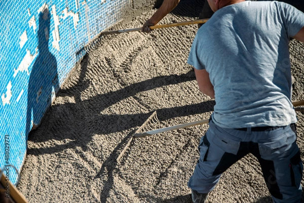 Obraz Workers in the sun spreading the concrete with rakes inside the pool, preparing the garden part