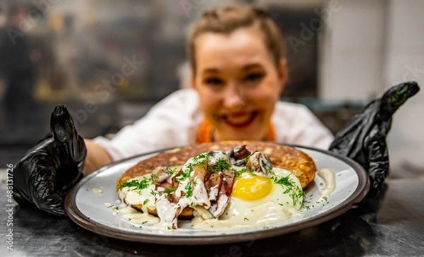 Fototapeta Chef's hands in gloves cooking fried pancake with mushroom sauce, bacon and egg on kitchen