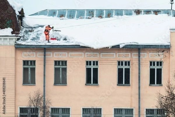 Fototapeta A working man in bright overalls with a safety belt with a shovel clears snow from the roof of an old building. Prevention of snow falling from the roof, industrial mountaineering