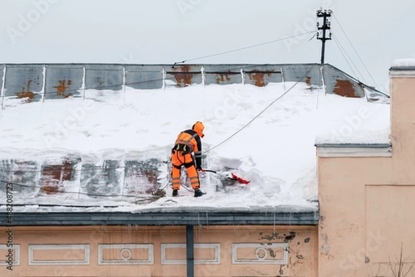 Fototapeta A working man in bright overalls with a safety belt with a shovel clears snow from the roof of an old building. Prevention of snow falling from the roof, industrial mountaineering