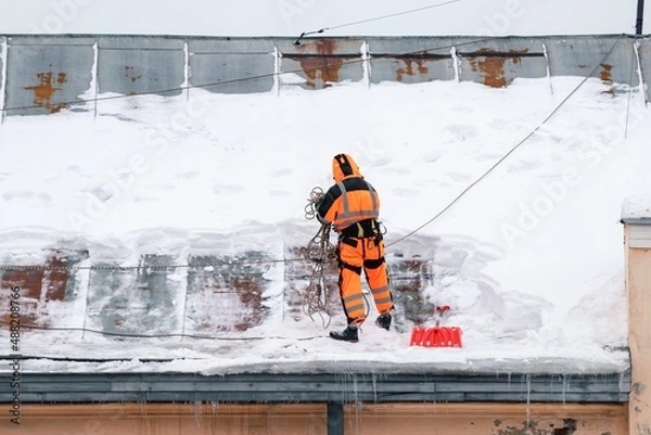 Fototapeta A working man in bright overalls with a safety belt with a shovel clears snow from the roof of an old building. Prevention of snow falling from the roof, industrial mountaineering