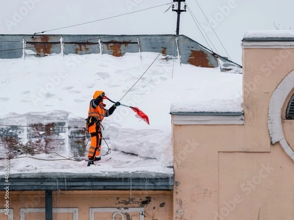 Fototapeta A working man in bright overalls with a safety belt with a shovel clears snow from the roof of an old building. Prevention of snow falling from the roof, industrial mountaineering