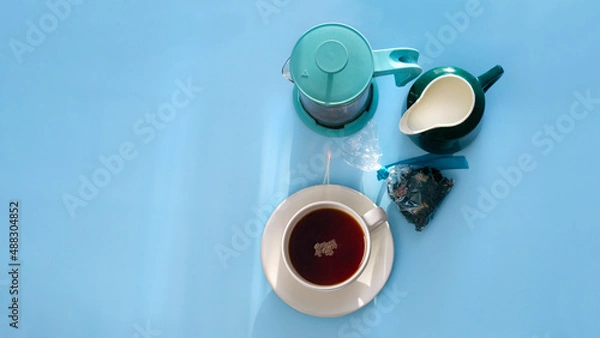 Fototapeta A cup of black hot tea with milk in jug and teapot stand on blue table with sunbeams. View from above.