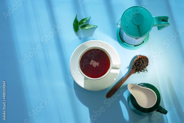 Fototapeta A cup of black hot tea with milk in jug and teapot stand on blue table with sunbeams. View from above.