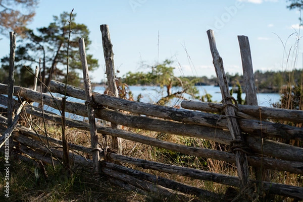 Fototapeta Traditional old wooden roundpole fence on the countryside. Idyllic view of the countryside and landscape with the water and forest in the background. Photo taken in Oskarshamn, Sweden.