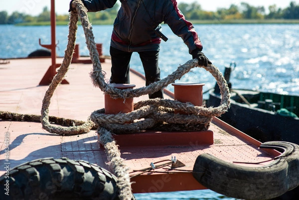 Fototapeta Sailor in overalls on the deck of the ship pulls the mooring cable in the port