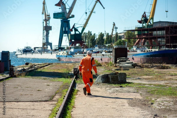 Fototapeta Shipyard worker in overalls against the background of cranes and ships