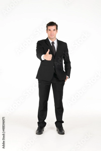 Fototapeta Portrait of a smiling western business man wearing a black suit with thumbs up and his hands in his pants pocket at studio shot on white background.