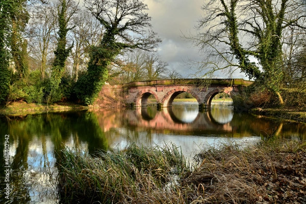 Obraz Eine schöne alte Sandsteinbrücke spiegelt sich im Wasser eines See im Frühling
in einer Parklandschaft
