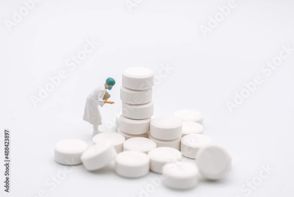 Obraz Healthcare and Medical Concept. Closeup of woman doctor miniature figure with patient file looking to stack of white pills on white background.