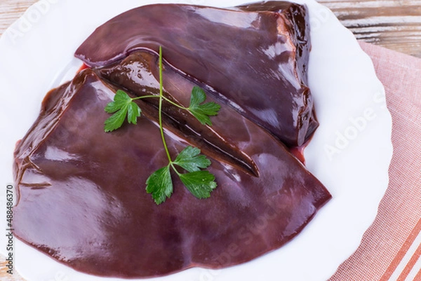 Fototapeta Raw liver on a white plate on a wooden background. Beef liver, top view. Offal in its raw form.