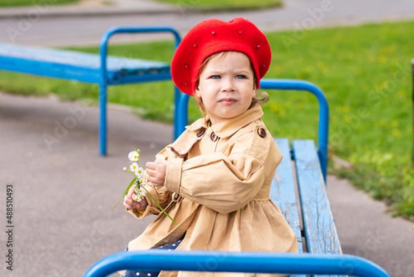 Obraz Charming Toddler in a beige raincoat and red beret admires a flower in spring.