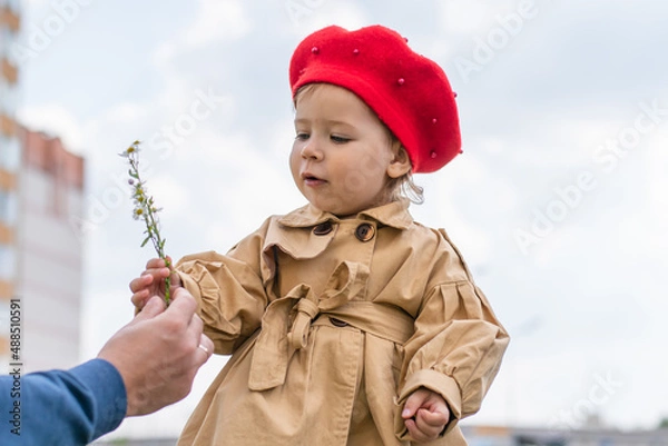 Obraz Charming Toddler in a beige raincoat and red beret admires a flower in spring.
