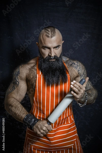 Fototapeta A brutal man with a long black beard in an apron holds a rolling pin in his hands