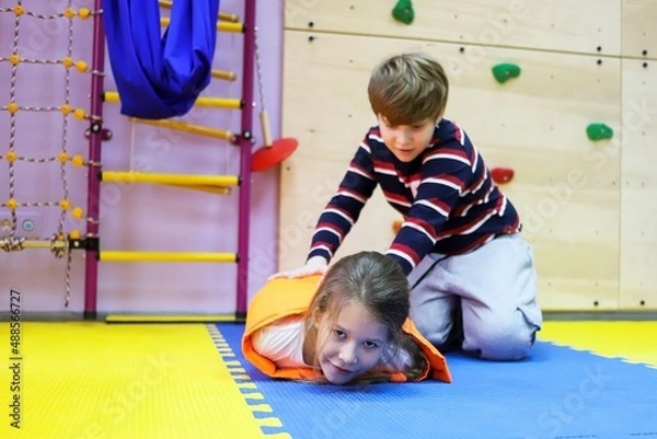 Fototapeta boy and girl children roll each other up in a thick blanket on mats controlling a small space for autism treatment in a correctional center