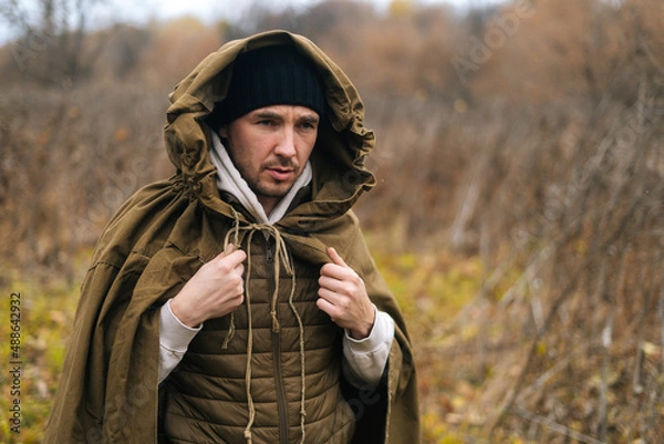 Fototapeta Portrait of thoughtful tourist man wearing green raincoat tent standing in thicket of bushes in cold overcast day and looking away. Concept of scout, research, travel and survival in nature.