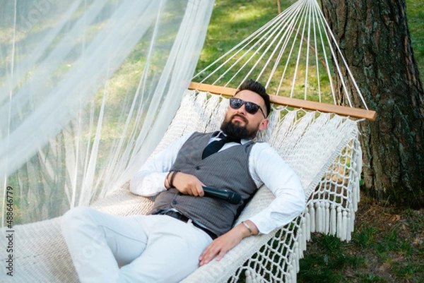 Fototapeta Bearded man in sunglasses and a suit  lies in a hammock outdoors. The host with the microphone is resting.