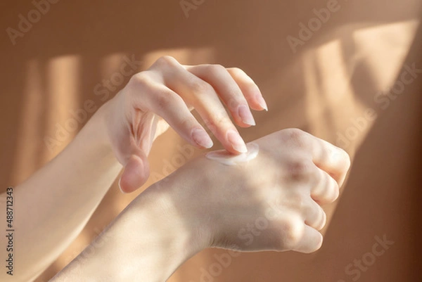 Obraz Application of cream, lotion on hands. Beautiful female hands rub moisturizing liquid into delicate skin, in rays of sunlight on brown background. Concept of care and beauty