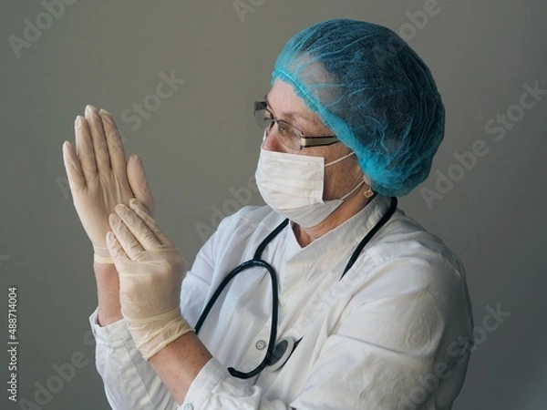Fototapeta Photo of an elderly woman doctor wearing a mask in uniform putting on gloves against a gray background. Profession saving lives. Help of older doctors during the coronavirus pandemic.