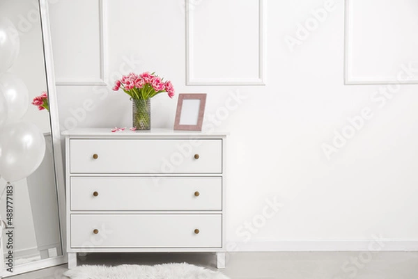 Fototapeta Vase with tulips and blank photo frame on chest of drawers near white wall in room
