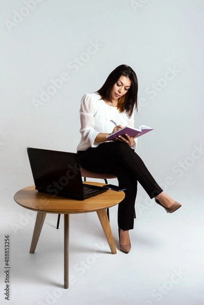 Fototapeta business woman posing while working at her desk. A laptop and some papers were placed in front of her. She is dressed in a strict black suit and talking on the phone. Isolated, white background.