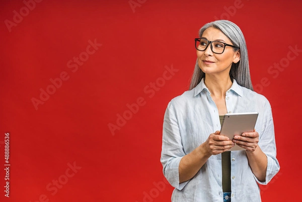 Fototapeta Portrait of smiling beautiful asian senior aged mature woman using tablet computer, isolated over red background. Copy space for text.