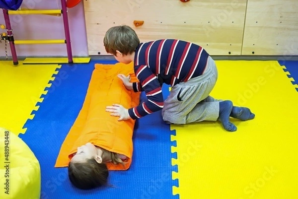 Fototapeta boy and girl children roll each other up in a thick blanket on mats controlling a small space for autism treatment in a correctional center