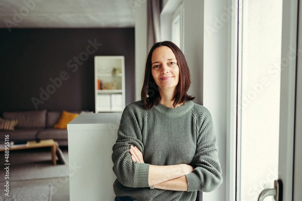 Obraz Portrait of smiling woman with arms crossed leaning by window at home