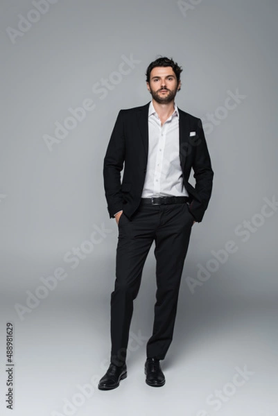 Fototapeta full length view of man in black elegant suit standing with hands in pockets while looking at camera on grey