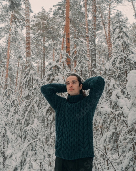 Fototapeta A young guy in a green sweater walks through a snowy forest