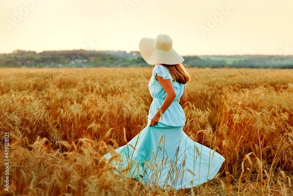 Fototapeta A young woman in a hat and a blue dress runs in a wheat field. The lady enjoys the sunset and nature.
