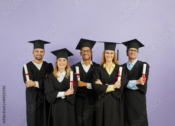 Fototapeta Portrait of group of happy multiracial university graduates with diplomas in their hands on purple background. Students dressed in mortarboards and in bachelor mantles stand in row and look at camera.