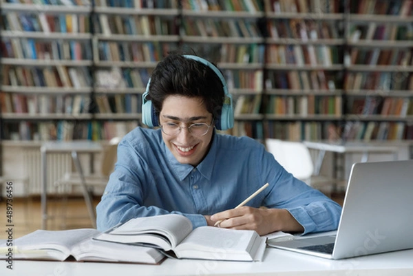Obraz Happy motivated millennial Jewish male student in glasses favorite music in headphones, feeling excited preparing for college exams, improving knowledge reading textbooks, sitting at table in library.