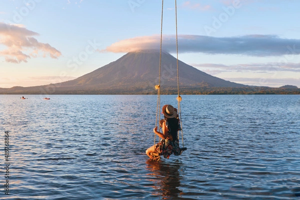Obraz Back view of woman sitting on a swing overlooking the volcano concenpcion on ometepe island, Nicaragua.