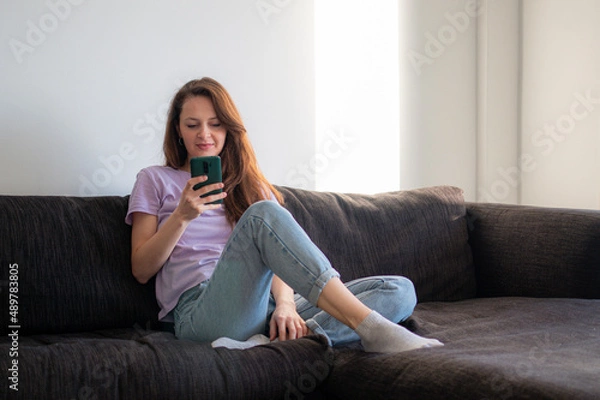 Fototapeta A young woman is sitting on a sofa and using her phone