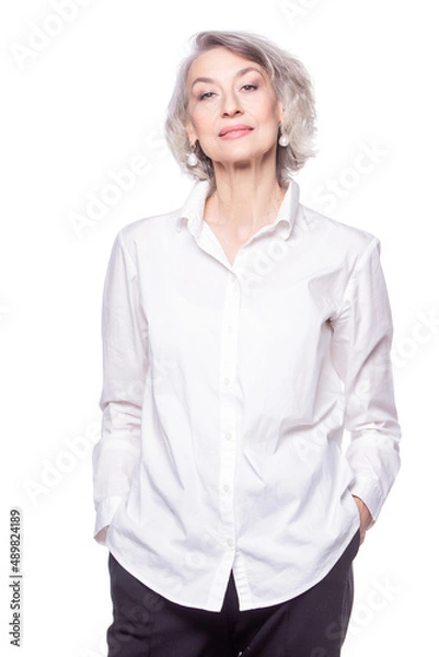 Fototapeta Happy trendy senior woman with a beautiful smile wearing a fashionable shirt standing with her hands in her pockets looking at the camera isolated on white background