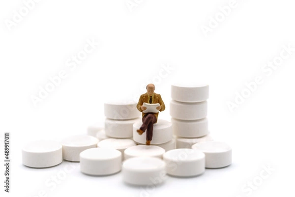 Obraz Healthcare, Medical and Information Concept. Closeup of old man businessman miniature figure sitting and reading book on stack of white pills on white background.